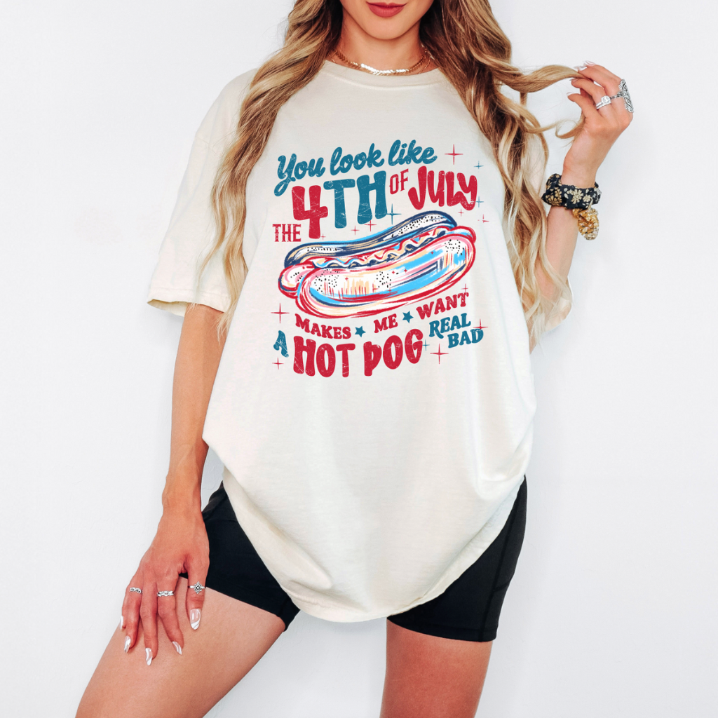 Woman wearing a cream oversized t-shirt with a red, white, and blue hot dog graphic and patriotic text that says &quot;You Look Like the 4th of July – Makes Me Want a Hot Dog Real Bad,&quot; styled with black biker shorts.