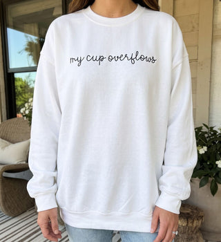 A person wearing a white crewneck sweatshirt with the phrase 'my cup overflows' printed in black cursive across the chest, paired with light blue jeans in an outdoor patio setting.