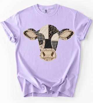 A light purple short-sleeve crewneck t-shirt featuring a large cow face patchwork graphic on the chest. The cow graphic is made of various patterned fabric sections including gingham, floral, and solid black and cream colors.