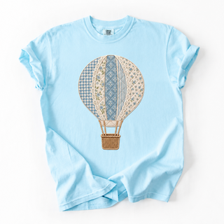 A light blue short-sleeve crewneck t-shirt featuring a central graphic of a vintage hot air balloon with a patchwork pattern of beige, blue, and brown fabrics, laid flat against a white background.