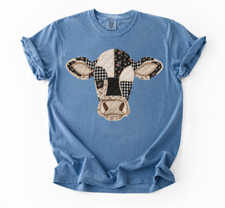 A light blue short-sleeve crewneck t-shirt featuring a cow head patchwork graphic on the front, laid flat against a white background.