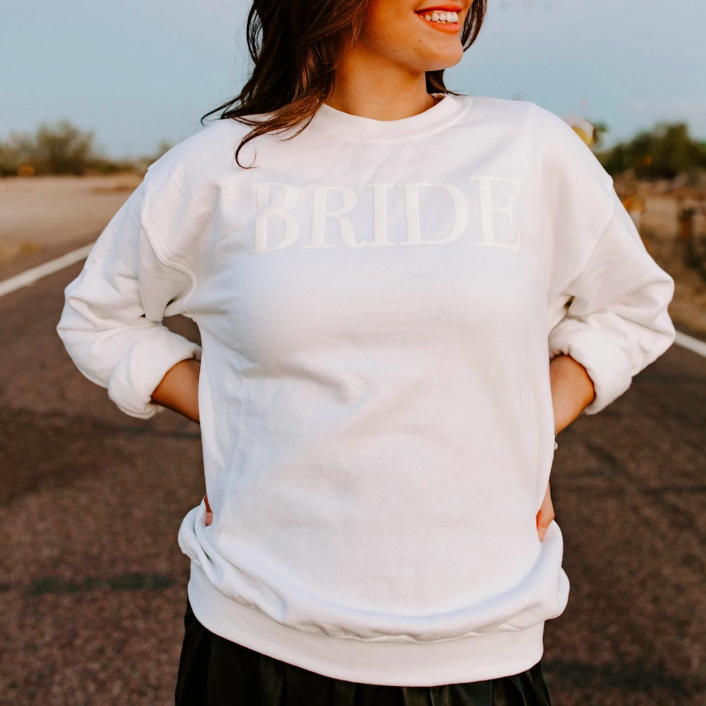 A women&#39;s crewneck sweatshirt with the word &quot;Bride&quot; elegantly embssed text on the front. This soft and comfortable white sweatshirt is perfect for the bride-to-be as she prepares for her big day. Available in unisex sizes Small to Extra Large, made from a preshrunk 50/50 cotton and polyester blend.