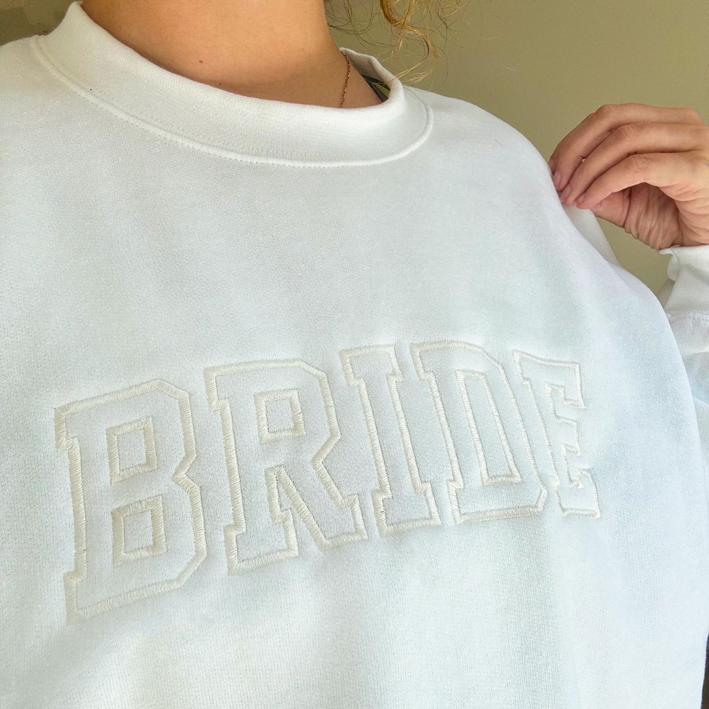 A women&#39;s embroidered crewneck sweatshirt with the word &quot;Bride&quot; elegantly stitched on the front. This soft and comfortable white sweatshirt is perfect for the bride-to-be as she prepares for her big day. Available in unisex sizes Small to Extra Large, made from a preshrunk 50/50 cotton and polyester blend.