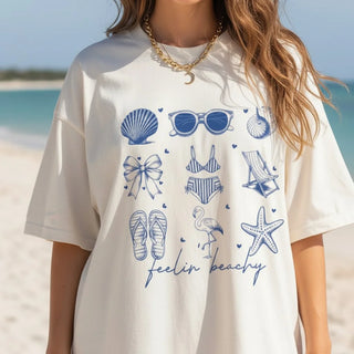 White t-shirt with blue beach-themed graphics and 'feelin' beachy' text on a beach background