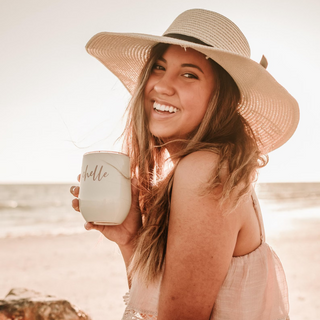 Woman on a beach holding a mug with 'hello' on it, wearing a wide-brimmed hat.