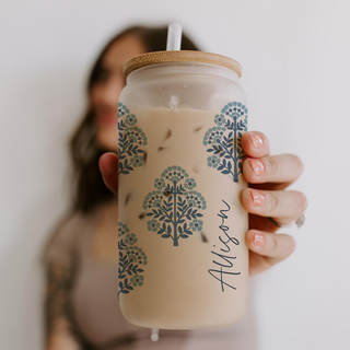 Person holding a glass with a floral design and wooden lid, blurred background