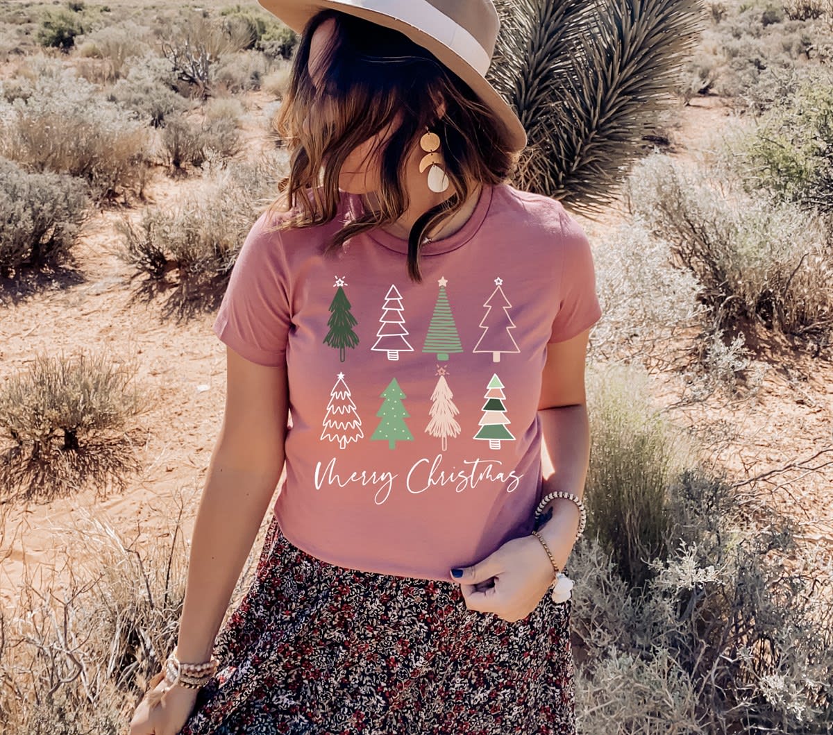 A women's graphic t-shirt with short sleeves, featuring Merry Christmas Trees design. The soft, comfortable tee comes in various colors like Heather Mauve and Heather Dust, and is available in sizes from Small to Extra Large.