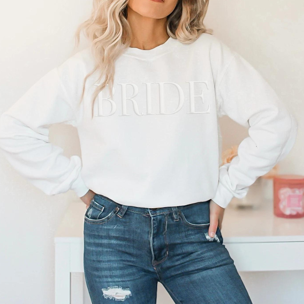 A women's crewneck sweatshirt with the word "Bride" elegantly embssed text on the front. This soft and comfortable white sweatshirt is perfect for the bride-to-be as she prepares for her big day. Available in unisex sizes Small to Extra Large, made from a preshrunk 50/50 cotton and polyester blend.